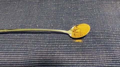 Ein Esslöffel mit gelbem Pulver liegt auf einem strukturierten blauen Tisch. Das Pulver könnte Hagebuttenpulver sein.