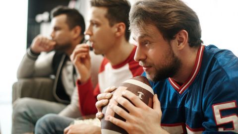 Drei Männer sitzen auf einer Couch, konzentriert auf ein Fußballspiel, während einer einen Football festhält.