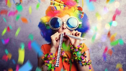 Eine Person mit bunter Perücke und großen Brillen bläst Luftschlangen inmitten von Konfetti bei nachhaltig Karneval feiern.