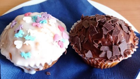 Zwei bunte Muffins liegen auf einem blauen Serviette: einer mit Zuckerguss und Streuseln, der andere mit Schokolade.
