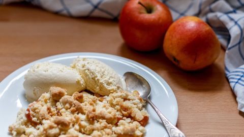 Auf einem Teller liegt ein warmes Apfelgericht mit Streuseln und Vanille-Eis, umgeben von frischen Äpfeln auf einem Holztisch.