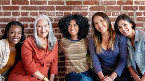 Fünf Frauen mit verschiedenen Hintergründen lächeln freundlich vor einer Backsteinwand, was Stärke und Gemeinschaft symbolisiert.