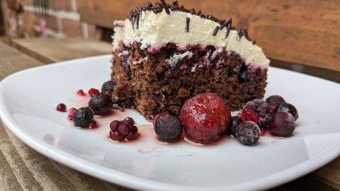 Eine saftige Schoko-Beerenschnitte liegt auf einem weißen Teller, umgeben von frischen, bunten Beeren und Fruchtsoße.