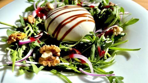 Ein Burrata auf frischem Rucola-Salat wird mit Walnüssen und roten Zwiebeln in einem hellen Teller serviert.