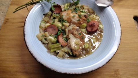 Eine Schüssel mit Reste-Suppe, gefüllt mit Gemüse, Hühnerteilen und Würstchen, steht auf einem Holzbrett.