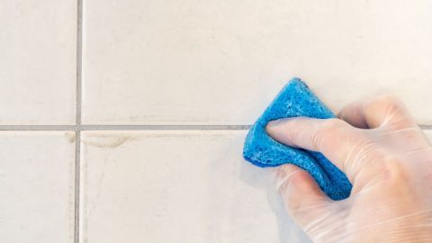 Ein blauer Schwamm reinigt die dunklen Fliesenfugen, um sie aufzuhellen. Der Hintergrund besteht aus hellen Fliesen.