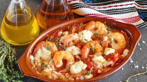 Garnelen liegen in Tomatensoße mit Feta-Käse in einer Schale, umgeben von Kräutern und Ölen auf einem dunklen Untergrund.