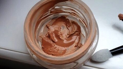 Ein Glas mit Make-Up in einem hellen Farbton steht auf einer Oberfläche und zeigt einen unbenutzten Schwamm daneben.