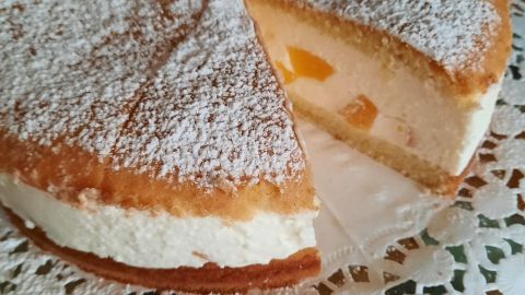 Eine Käse-Sahne-Torte mit Pfirsichen, bestäubt mit Puderzucker, liegt auf einer Spitzenunterlage, während ein Stück herausgeschnitten wurde.