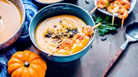 Eine cremige Kürbissuppe in einer Schüssel, garniert mit Garnelen und Kräutern, umgeben von Kürbissen und Besteck.