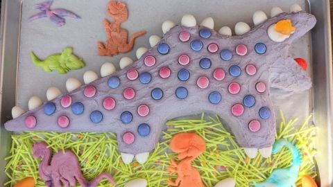 Ein großer, dekorativer Dino-Kuchen liegt auf einem Blech, umgeben von bunten Zuckerfiguren und Gras-Deko.