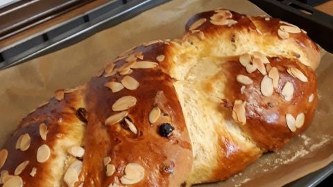 Ein goldbrauner Osterzopf mit Rosinen und Mandelsplittern ruht auf einem Backblech in einer modernen Küche.