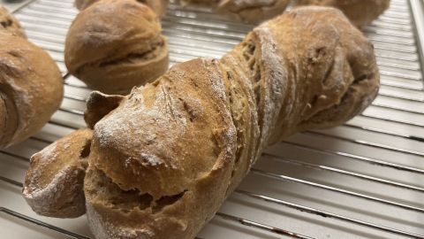 Ein goldenes Wurzelbrot aus Roggensauerteig ruht auf einem Gitter, um abzukühlen, umgeben von weiteren Brötchen.