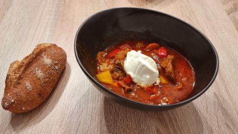 Eine Schüssel Gulaschsuppe mit buntem Gemüse und einem Klecks Sahne steht auf einem Tisch neben einem Brötchen.