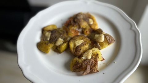 Goldbraun gebratene Quetschkartoffeln liegen auf einem weißen Teller, garniert mit einer hellen Sauce und serviert im Innenraum.