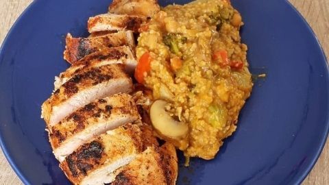 Gedämpfter Gemüsecouscous liegt neben knusprig gegrillten Hähnchenbrustscheiben auf einem blauen Teller.