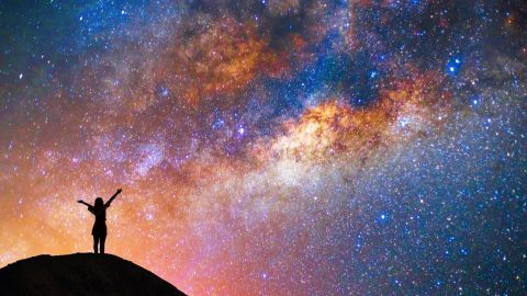 Eine Person steht auf einem Felsen und breitet die Arme aus, während sie das Universum mit Sterne und Galaxien bewundert.