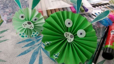Zwei niedliche Papier-Osterhasen stehen auf einem Tisch, während Bastelmaterialien und Farben im Hintergrund liegen.