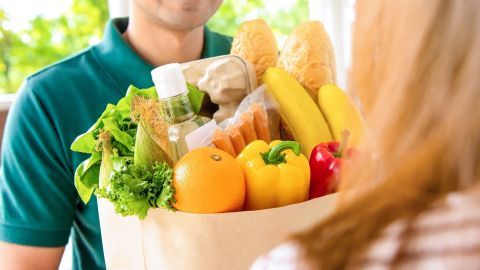 Ein Mann hält eine Tüte voller frischer Zutaten wie Gemüse, Obst und Brot, während er jemandem die Lieferung überreicht.