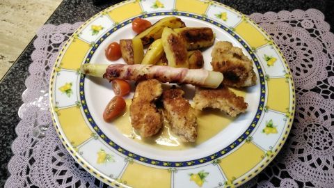 Frittierte Chicken-Nuggets liegen auf einem bunten Teller, umgeben von Spargel im Speckmantel und Kartoffeln.