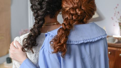 Zwei Frauen mit geflochtenem Haar stehen Rücken an Rücken, umgeben von einem hellen Raum mit Holzmöbeln.