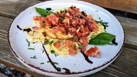 Ein saftiges Hähnchenschnitzel liegt auf Nudeln, garniert mit Bruschetta und frischen Basilikumblättern, auf einem Holzstahltisch.