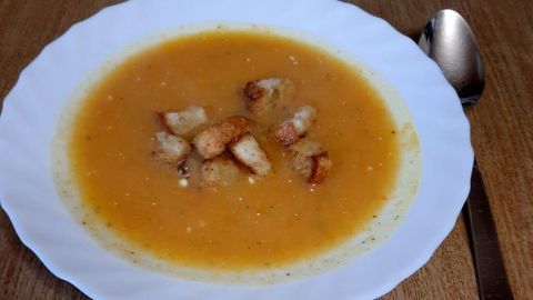 Eine cremige Wurzelgemüse-Suppe in einem weißen Teller, garniert mit knusprigen Brotwürfeln, steht auf einem Holztisch.