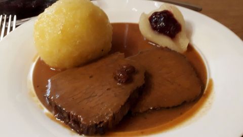 Zart gekochter Sauerbraten liegt auf einem Teller, umgeben von einem Kloß und Apfelkompott in aromatischer Soße.