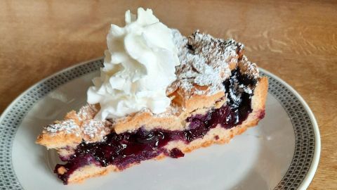 Ein Stück Gitterkuchen mit Heidelbeeren und Sahne steht auf einem Teller, umgeben von einer Holzoberfläche.