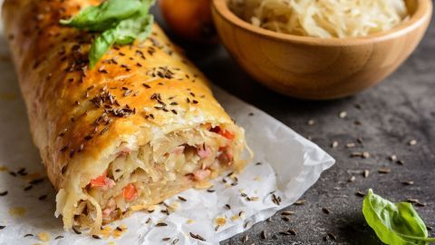 Ein goldbraun gebackener Sauerkrautstrudel liegt auf Backpapier, umgeben von frischem Kraut und einer Schüssel.
