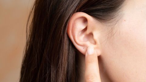 Eine Person berührt mit dem Finger ihr Ohr, was auf ein mögliches Jucken hinweist. Der Hintergrund ist neutral.