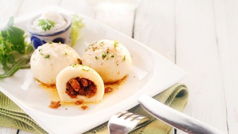 Auf dem Teller liegen drei Grammelknödel aus Kartoffelteig, einer ist aufgeschnitten und zeigt die Füllung. Daneben steht ein kleiner Schälchen mit einer weißen Beilage, umgeben von frischen Kräutern.