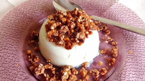 Ein cremiges Käse-Sahne-Dessert ist auf einem lila Teller mit Mandelkrokant dekoriert, umgeben von einem weißen Untergrund.