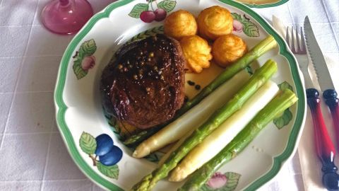 Auf einem Teller liegen zarte Rinderfilets, umgeben von grünem und weißem Spargel sowie kleinen Beilagen, alles auf einem hübschen Tisch gedeckt.