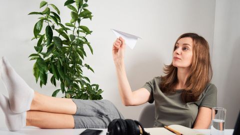 Eine Frau mit ausgestreckten Beinen sitzt entspannt am Schreibtisch und wirft ein Papierflugzeug, um Aufschieberitis zu bekämpfen.