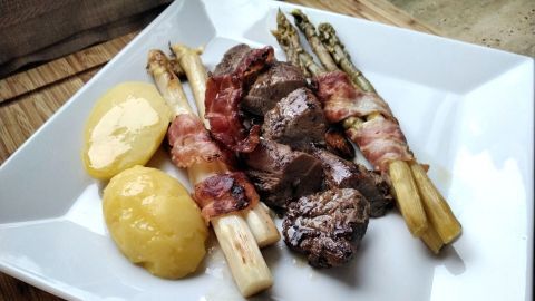 Lammfilet und mit Speck umwickelter Spargel liegen appetitlich auf einem weißen Teller, ergänzt von Pellkartoffeln.