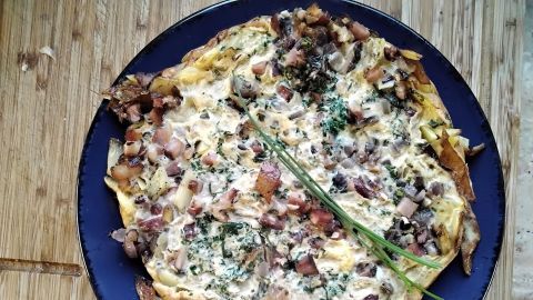 Ein Omelette mit Kartoffeln und Speck liegt auf einem blauen Teller, garniert mit frischen Kräutern, auf einem Holztisch.