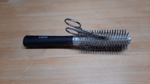Eine Rundbürste mit Haaren und einer Nagelschere liegt auf einem hellen Holztisch.
