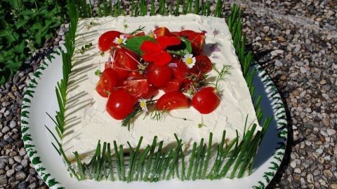 Eine üppige Sandwichtorte mit Frischkäse, dekoriert mit Tomaten, Kräutern und Gemüse, auf einem Teller im Freien.