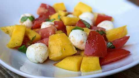 Bunte Würfel aus Mango und Tomaten liegen zusammen mit Mozzarella-Kugeln auf einem weißen Teller, garniert mit frischem Basilikum.