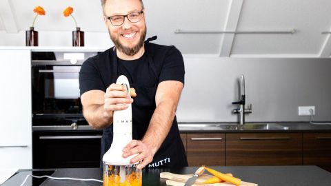 Ein Mann mit Brille und Schürze benutzt einen Mixer, um frische Zutaten in einer modernen Küche zu pürieren.