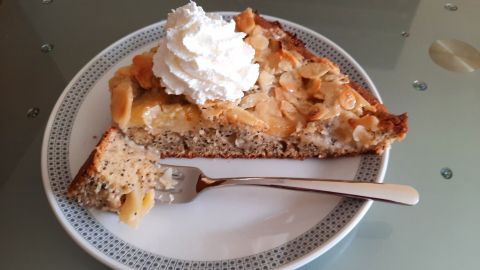 Eine Stück Apfel-Mohn-Kuchen mit Sahnehaube liegt auf einem Teller, umgeben von einem Tisch mit Glasoberfläche.