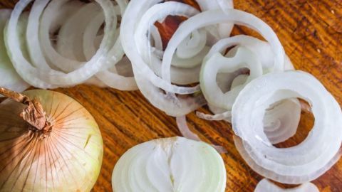 Eine Zwiebel liegt halbiert und ungeschnitten auf einem Holzbrett, während feine Zwiebelringe daneben verteilt sind.