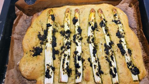 Die goldbraune Focaccia hat grüne Bärlauchpaste und frischen Spargel, auf einem Blech in einer Küchenumgebung.