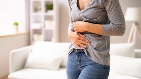 Eine Person mit Bauchschmerzen hält sich den Bauch in einem hellen Raum mit einem Sofa, was auf Sodbrennen oder Gastritis hindeutet.