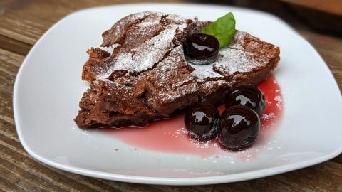 Ein Stück schokoladiger Brownie liegt auf einem weißen Teller, garniert mit Kirschen und Puderzucker.