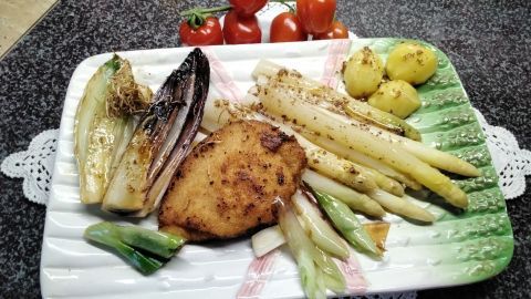Weißer Spargel liegt auf einem Teller, umgeben von Bratkartoffeln und einer panierten Frikadelle, mit Kirschtomaten im Hintergrund.