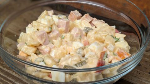 Ein bunter Kartoffelsalat mit Fleischwurst und Apfel wird in einer Glasschüssel serviert, umgeben von einer rustikalen Holzoberfläche.