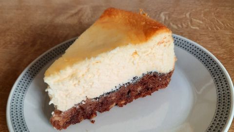 Ein Stück Käse-Schmand-Kuchen liegt auf einem Teller, der Mohn-Keks-Boden ist deutlich sichtbar und umgeben von Holz.