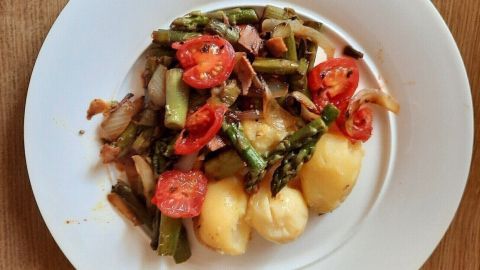 Auf einem weißen Teller liegen grüne Spargel und Kartoffeln in Knoblauchbutter, umgeben von frischen Tomaten und Zwiebeln.